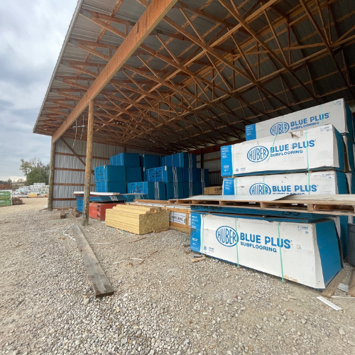 Open-sided warehouse with stacks of Huber Blue Plus subflooring boxes and wooden planks on gravel. Cloudy sky adds a neutral tone to the scene.