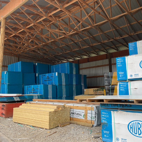 Inside a large storage building, stacks of blue and white insulated panels and pallets of wooden beams are neatly arranged, creating an orderly space.
