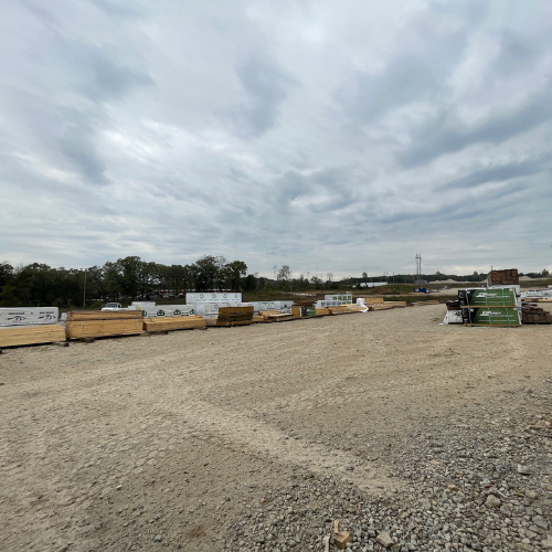 A construction site with stacks of lumber and building materials under a cloudy sky. Trees and utility poles are visible in the background.