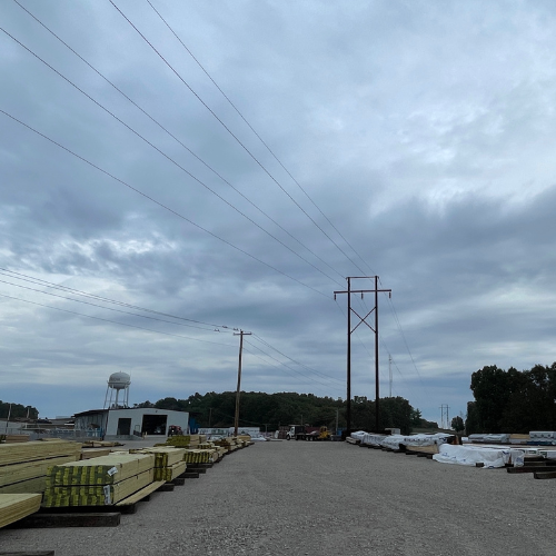 Gravel lot with stacked lumber and covered materials, power lines stretching across a cloudy sky, a water tower, and trees in the distance. Overcast mood.