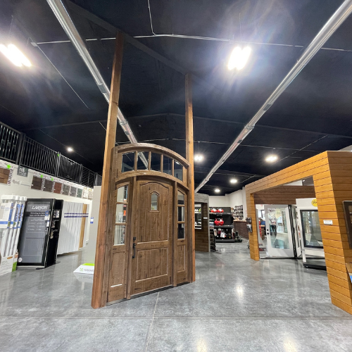 A showroom interior displaying a wooden entry door with an arched window at the top, set against a sleek, polished concrete floor and surrounded by modern lighting.