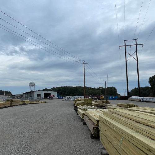 Lumber yard under a cloudy sky with stacked wooden planks and utility poles. Buildings and trees in the distance provide an industrial setting.