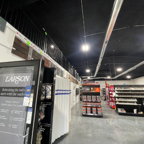 Hardware store interior with a high ceiling, featuring neatly arranged shelves of products, a display door, and bright fluorescent lighting. The atmosphere is organized and welcoming.