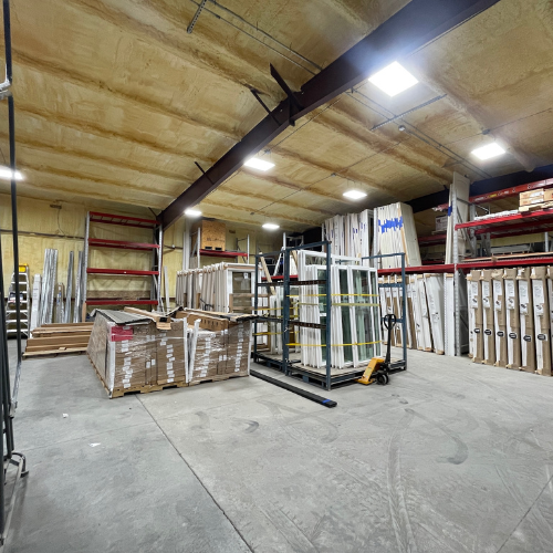 Warehouse interior with high ceilings, lit by bright overhead lights. Stacked windows, pallets, and shelving units fill the spacious, organized area.