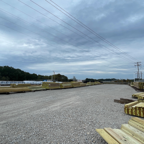 A gravel yard with stacks of wooden planks organized neatly under an overcast sky. Power lines run across, and trees line the horizon, creating an industrial and serene scene.