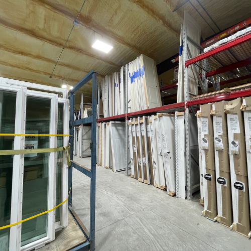 Spacious warehouse with organized stacks of doors and windows on metal shelves. Bright lighting illuminates the high ceiling, creating an orderly atmosphere.