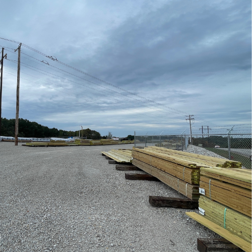 Stacks of wooden planks are neatly arranged in a gravel lot near a chain-link fence. Overcast skies and power lines enhance the industrial setting.