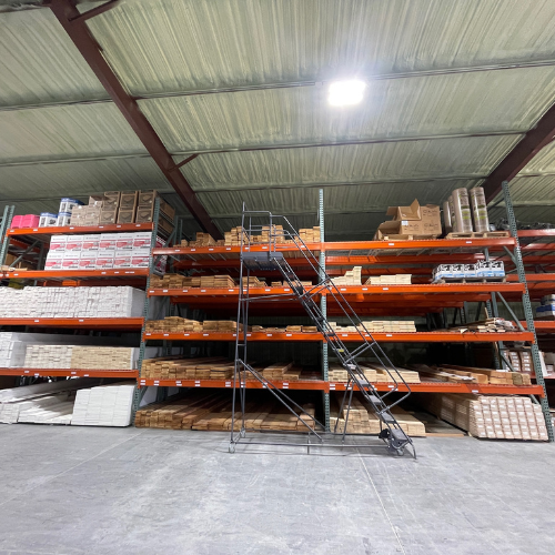 Warehouse interior with tall metal shelves stocked with various packages and boxes. A metal rolling ladder is positioned in front, under bright lighting.
