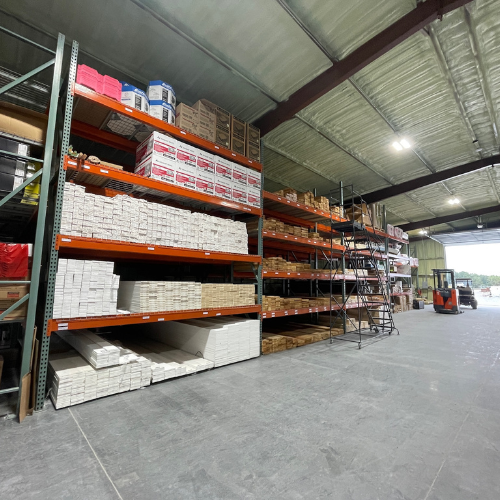 Spacious warehouse with tall shelves stacked with various boxes and materials. A ladder and forklift are seen on the right, suggesting organized stock management.