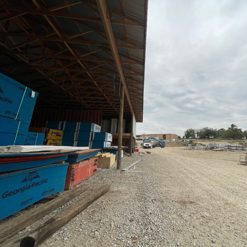 A gravel lot with stacks of blue and red lumber under a metal-roofed structure. Overcast sky and distant buildings suggest a construction site.