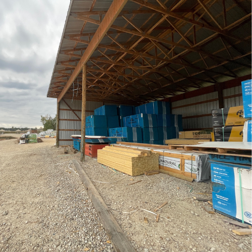 Outdoor storage area with an open-roof shelter housing various building materials, including stacked blue and yellow insulation boards, on a gravel lot. Cloudy sky.
