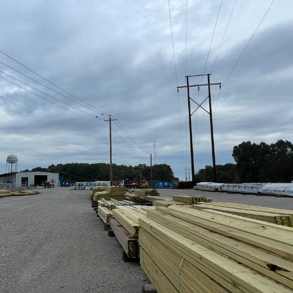 LumberLumber yard under cloudy skies with stacked wooden beams and utility poles. Industrial feel with distant trees and storage buildings.