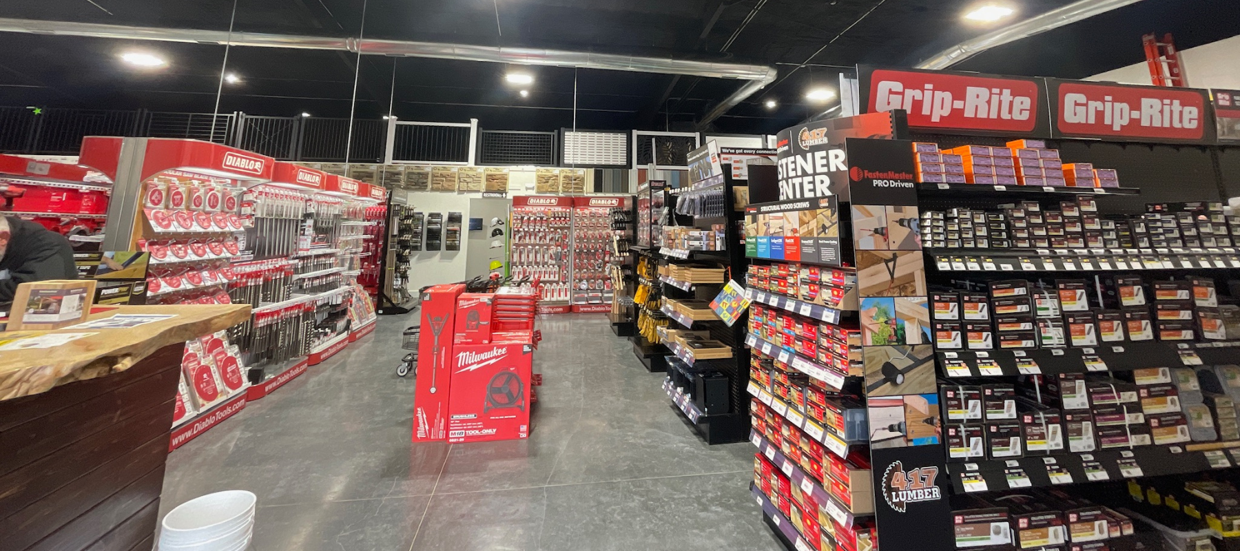A hardware store aisle with bright lighting, featuring red Milwaukee tool displays on the left and Grip-Rite fasteners on the right, creating an organized and professional atmosphere.